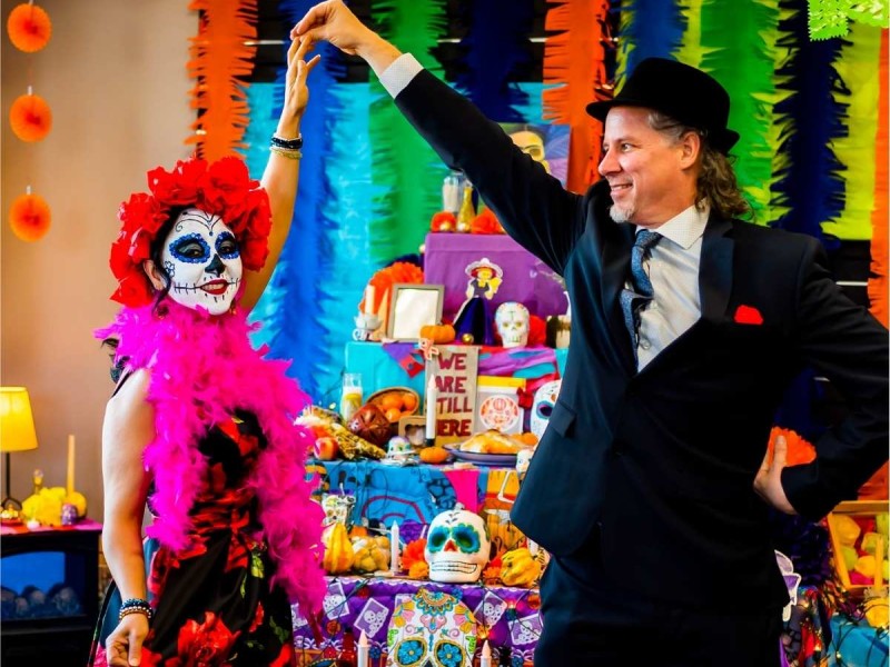 A woman dressed in regalia for Day of the Dead stands alongside with a man dressed in a black suit and hat