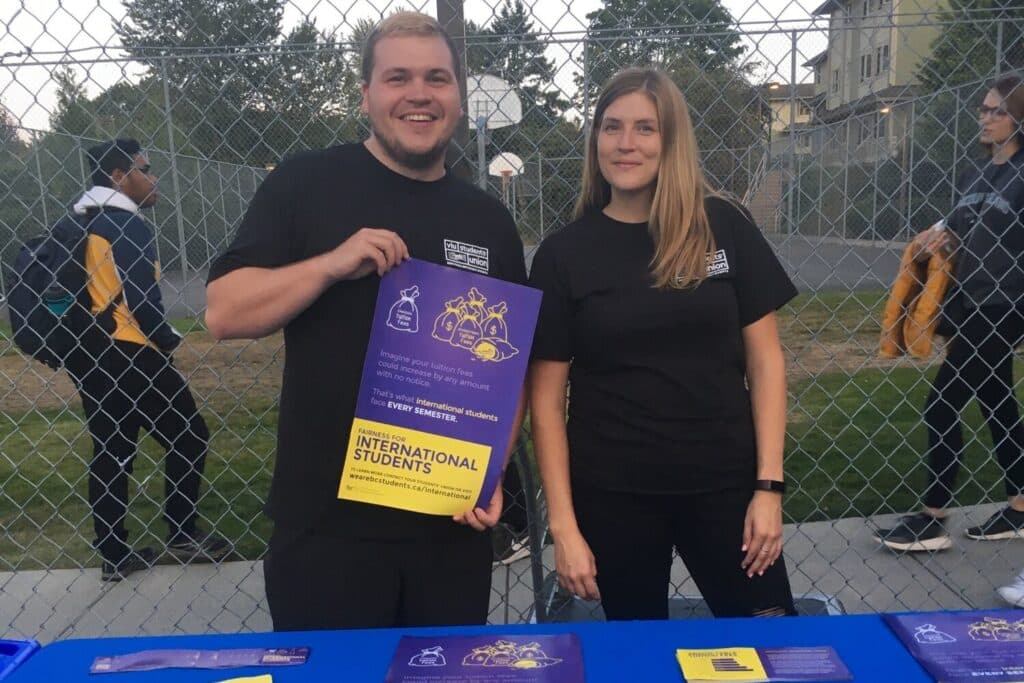 Two people stand in front of a booth and hold up a poster.
